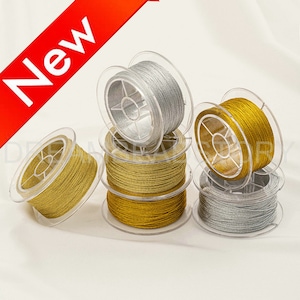 Könnte beinhalten: Sechs Spulen mit Metallic-Garn, drei goldfarben und drei silberfarben, liegen auf einer weißen Oberfläche. Die Spulen sind aus durchsichtigem Kunststoff mit einer zentralen Spindel.