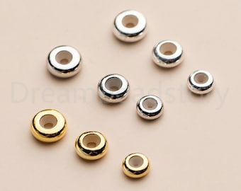 May include: A selection of silver and gold-coloured round beads with a hole through the centre. The beads vary in size and are scattered on a light beige surface. The metallic beads have a shiny finish.