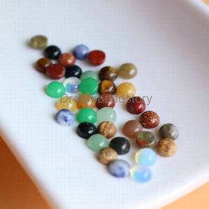 May include: Assortment of colorful round gemstone cabochons in various colors, including green, blue, red, brown, and yellow. The cabochons are arranged on a white surface.