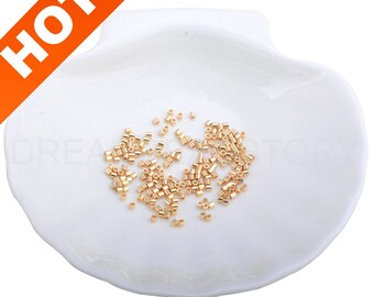 May include: A white ceramic dish with a shell-like shape holds a pile of small, gold-coloured metal jump rings. The jump rings are about 3mm in diameter.