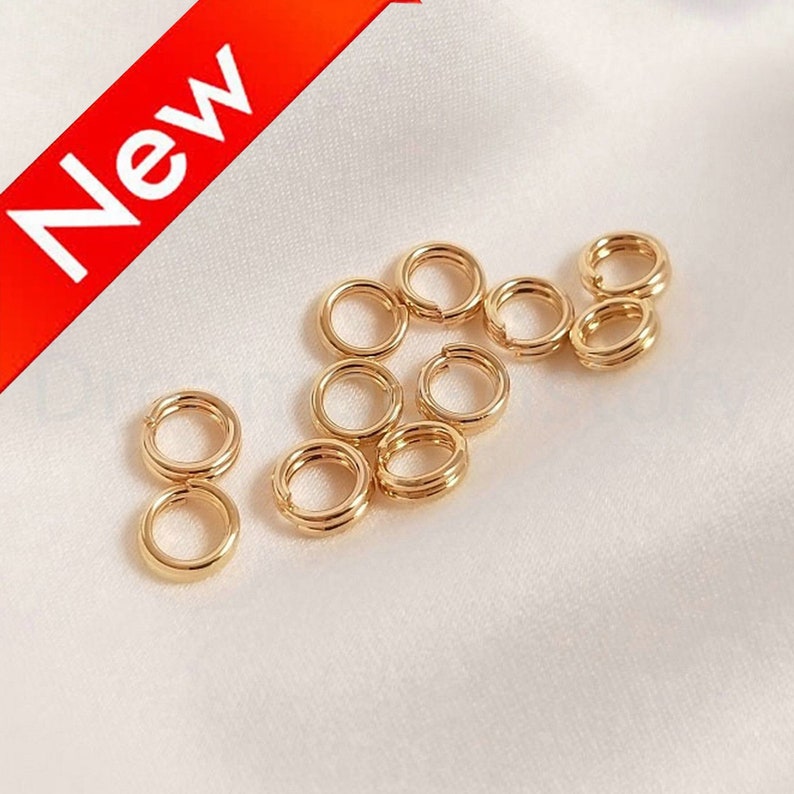 以下が含まれることがあります： 白い布の表面に、12個の金色のジャンプリング（別名スプリットリング）が散らばっています。これらのリングは、ジュエリー作りやクラフトに使用されます。