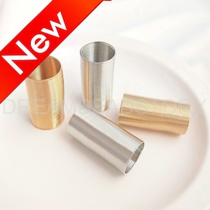 Puede incluir: Cuatro carretes de alambre, dos dorados y dos plateados, están dispuestos sobre una superficie blanca. Los carretes están hechos de alambre metálico fino que está enrollado apretado en una bobina.