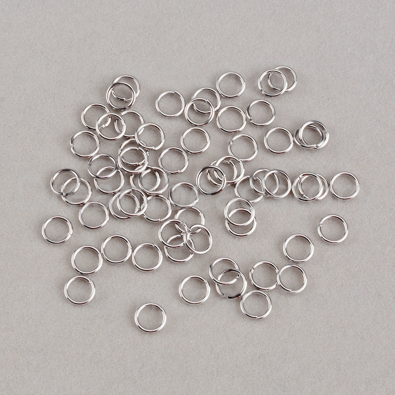 May include: A large quantity of silver jump rings, scattered on a gray surface.