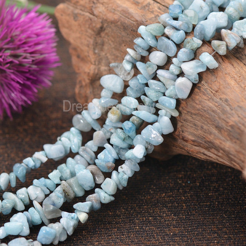 May include: A strand of light blue aquamarine chip beads. The beads are irregular in shape and have a natural, rough texture. The beads are strung on a clear thread and are displayed on a piece of wood.