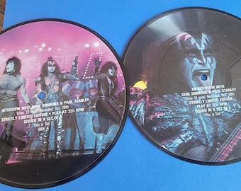 May include: Two circular vinyl picture discs featuring the band KISS. The left disc shows the band members on stage with text reading An Interview with Gene Simmons & Paul Stanley. The right disc features a close-up of Gene Simmons with text.