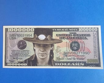 May include: A novelty one million dollar bill featuring a portrait of Stevie Ray Vaughan wearing a wide-brimmed hat. The note includes the text United States of America, Blues Hall of Fame, Couldn't Stand The Weather, and SRV10031954.
