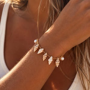 Peut inclure: Un bracelet à chaîne dorée orné d'une série de petits coquillages en spirale, de couleur beige et blanche, alternés avec des perles rondes, blanches et lustrées. Les maillons métalliques délicats relient chaque breloque en motif répété.