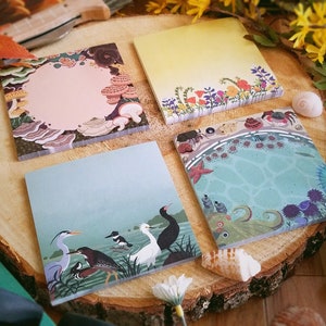 May include: Four square notepads with nature-themed illustrations. One features mushrooms, another flowers, a third birds, and the last an underwater scene with an octopus and starfish. The pads are arranged on a wooden slice.