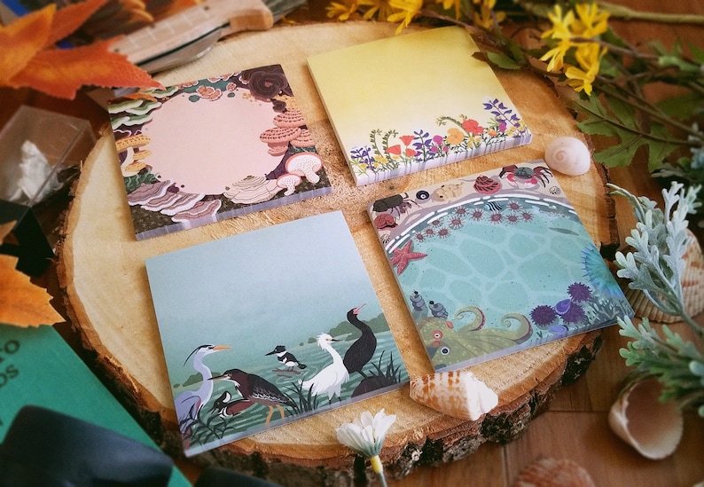 May include: Four square notepads with nature-themed illustrations. One features mushrooms, another flowers, a third birds, and the last an underwater scene with an octopus and starfish. The pads are arranged on a wooden slice.