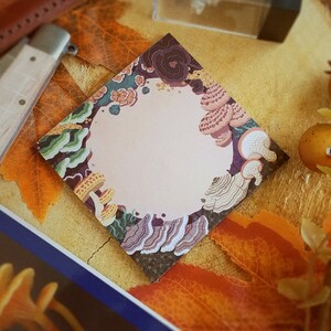 May include: A square notepad with a light pink center, surrounded by a border of colorful mushrooms and fungi. The design features shades of brown, green, and yellow, with a diamond-shaped layout. The notepad is placed on a surface with autumn leaves.