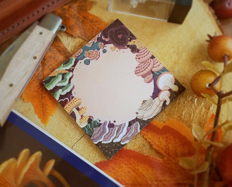 May include: A square notepad with a light pink center, surrounded by a border of colorful mushrooms and fungi. The design features shades of brown, green, and yellow, with a diamond-shaped layout. The notepad is placed on a surface with autumn leaves.