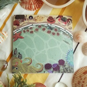 May include: A colorful illustrated notepad with an octopus, sea anemones, starfish, and crabs on a green ocean background. The notepad is on a striped yellow and white surface.