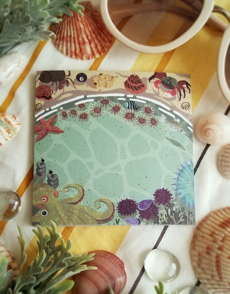 May include: A colorful illustrated notepad with an octopus, sea anemones, starfish, and crabs on a green ocean background. The notepad is on a striped yellow and white surface.
