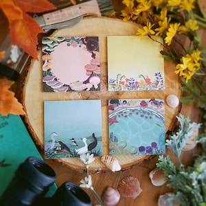 May include: Four square notepads with colorful illustrations are arranged on a wooden surface. The pads feature designs of mushrooms, flowers, birds, and an underwater scene. The pads are surrounded by flowers, leaves, and seashells.