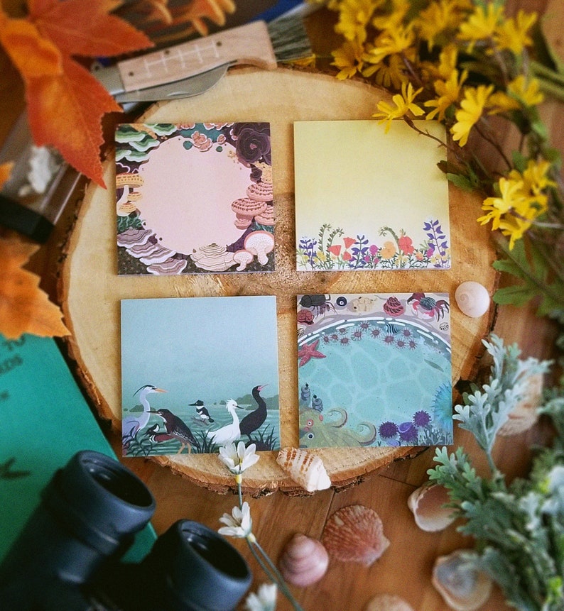May include: Four square notepads with colorful illustrations are arranged on a wooden surface. The pads feature designs of mushrooms, flowers, birds, and an underwater scene. The pads are surrounded by flowers, leaves, and seashells.