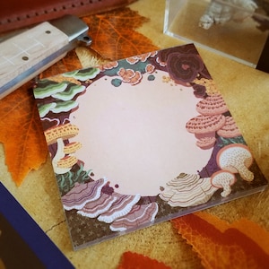May include: Square notepad with a mushroom illustration border. The design features various mushroom types in shades of brown, purple, and green, surrounding a blank, light pink center. The notepad is on a wooden surface with fall leaves.