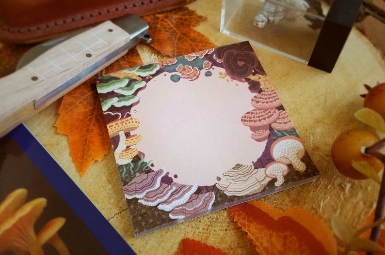 May include: Square notepad with a mushroom illustration border. The design features various mushroom types in shades of brown, purple, and green, surrounding a blank, light pink center. The notepad is on a wooden surface with fall leaves.