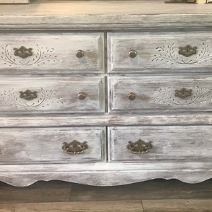 May include: A distressed white six-drawer chest of drawers with ornate drawer pulls and carved floral details. The chest has a weathered, vintage appearance, with a curved base and brass hardware.