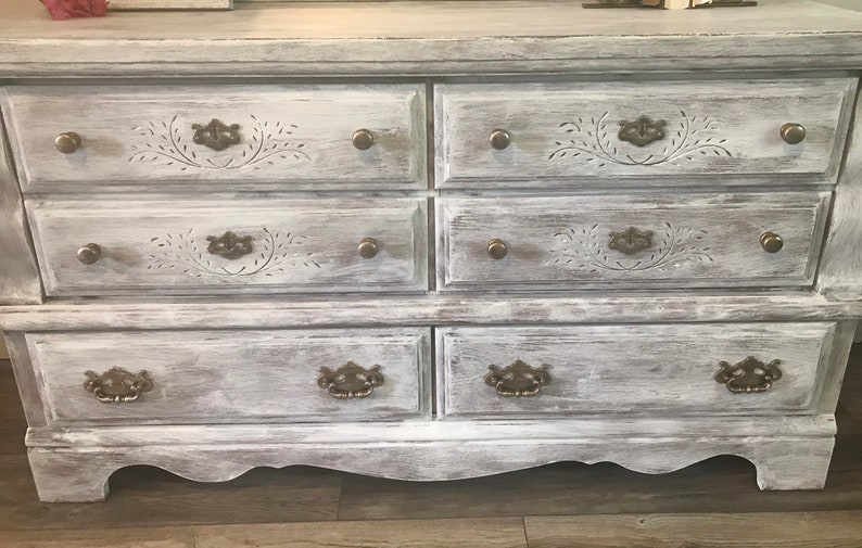 May include: A distressed white six-drawer chest of drawers with ornate drawer pulls and carved floral details. The chest has a weathered, vintage appearance, with a curved base and brass hardware.