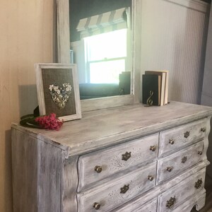 May include: A distressed white dresser with a matching mirror. The dresser has seven drawers with ornate brass hardware. A framed heart-shaped floral arrangement and a small stack of books sit on top. Pink flowers are placed on the dresser.