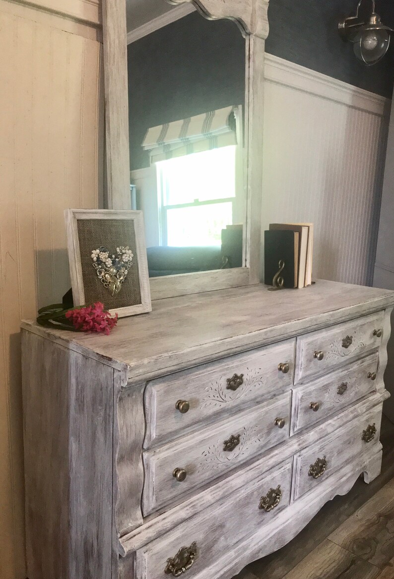 May include: A distressed white dresser with a matching mirror. The dresser has seven drawers with ornate brass hardware. A framed heart-shaped floral arrangement and a small stack of books sit on top. Pink flowers are placed on the dresser.