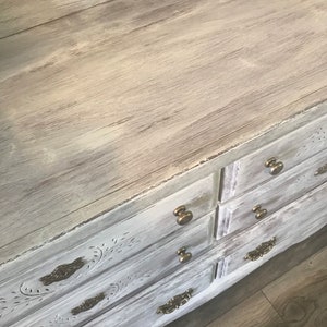 May include: A distressed white chest of drawers with six drawers and brass handles. The top surface has a light wood grain effect. The drawer fronts feature carved floral designs, creating a vintage aesthetic.