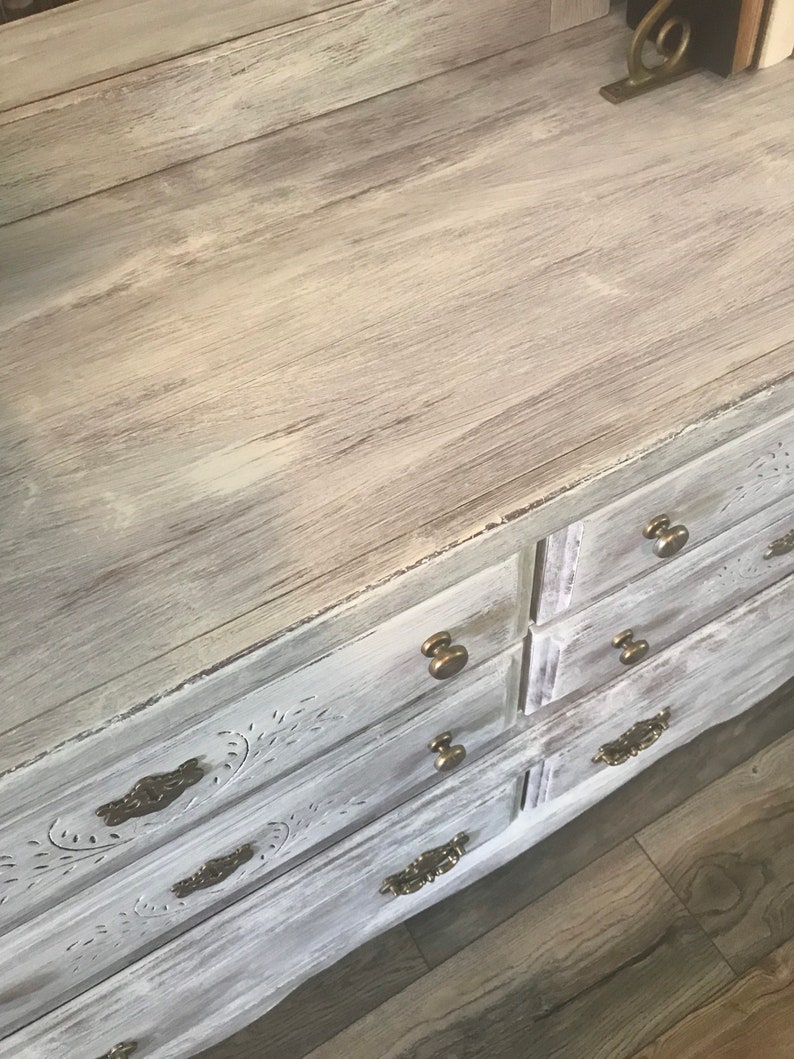 May include: A distressed white chest of drawers with six drawers and brass handles. The top surface has a light wood grain effect. The drawer fronts feature carved floral designs, creating a vintage aesthetic.