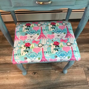 May include: A light blue vanity table with a drawer and a decorative stool. The stool's seat features a colorful pattern with the Eiffel Tower, butterflies, hearts, and the word "Paris". The table and stool are on a wooden floor.