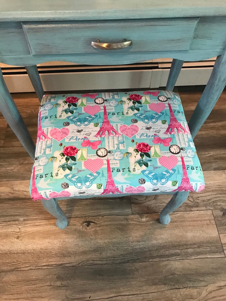 May include: A light blue vanity table with a drawer and a decorative stool. The stool's seat features a colorful pattern with the Eiffel Tower, butterflies, hearts, and the word "Paris". The table and stool are on a wooden floor.