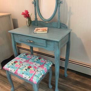 May include: A vintage-style vanity set in a distressed light blue finish. The set includes a small table with a drawer, an oval mirror, and a matching stool with a Paris-themed fabric seat. A small glass jar with pink flowers sits on the table.