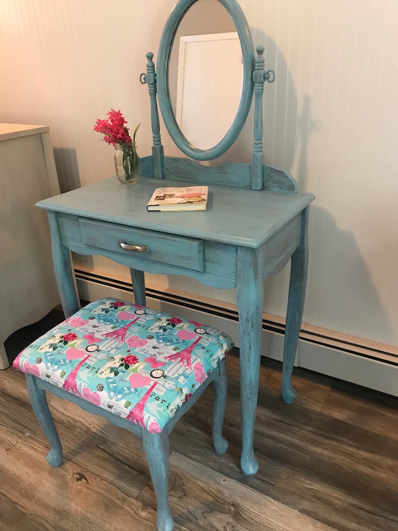 May include: A vintage-style vanity set in a distressed light blue finish. The set includes a small table with a drawer, an oval mirror, and a matching stool with a Paris-themed fabric seat. A small glass jar with pink flowers sits on the table.