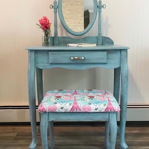May include: A vintage-style vanity table and stool set in a distressed teal finish. The oval mirror is attached to the table. The stool has a patterned seat with Eiffel Towers and hearts. A small drawer is visible.