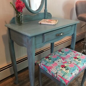 May include: A vintage-style vanity table and stool set painted in a distressed teal finish. The vanity features a round mirror, a drawer, and a floral arrangement. The stool has a patterned seat with Eiffel Towers and roses.