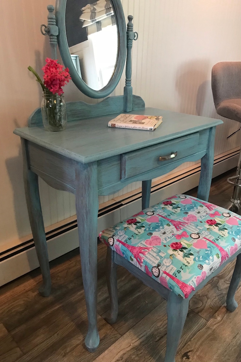 May include: A vintage-style vanity table and stool set painted in a distressed teal finish. The vanity features a round mirror, a drawer, and a floral arrangement. The stool has a patterned seat with Eiffel Towers and roses.