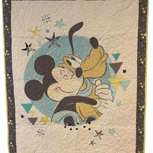 May include: A quilted baby blanket featuring Mickey Mouse and Pluto embracing within a light blue circle. The blanket has a cream background with star and triangle accents, and a patterned border in gray and yellow.