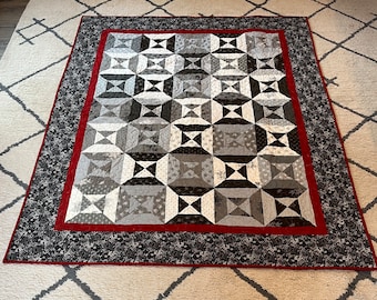 May include: A quilt with a geometric pattern in shades of gray and white, framed by a black and red border.