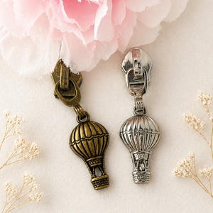 May include: Two decorative zipper pulls with hot air balloon charms. One pull is antique brass, the other is silver. The charms are detailed with a basket and balloon design. The background includes a pink flower and small white flowers.