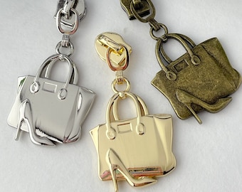 Könnte beinhalten: Drei Reißverschlussanhänger in Form einer Handtasche mit einem hohen Absatzschuh. Die Reißverschlussanhänger sind silber, gold und bronzefarben.