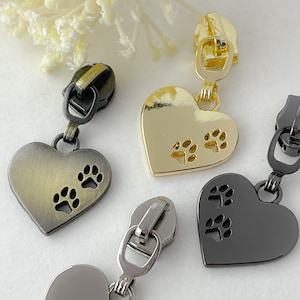May include: Four zipper pulls with heart-shaped charms featuring paw print cutouts. The charms are in gold, silver, black, and antique gold.