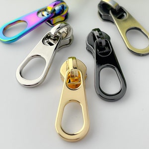 May include: Four zipper pulls in different colors and finishes: iridescent, silver, gold, and black.