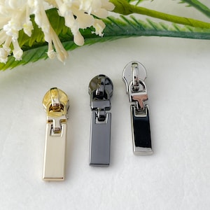 May include: Three zipper pulls in different finishes: gold, black, and silver. Each pull has a rectangular slider and a small loop at the top.