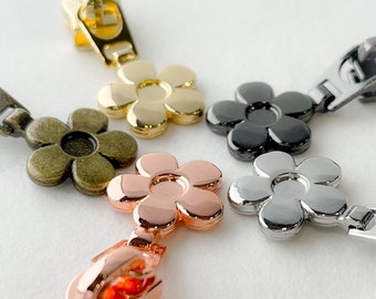 May include: Five zipper pulls in different colors: gold, black, bronze, rose gold, and silver. Each pull is shaped like a daisy.