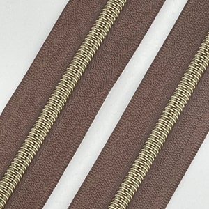 May include: Two brown zipper tapes with gold metal teeth. The zipper tapes are laid out side by side.
