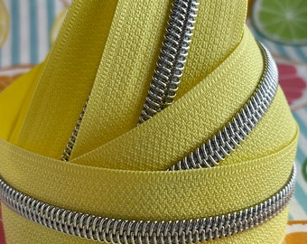 May include: Close-up of a yellow zipper tape with a silver metal coil. The zipper tape is folded in a spiral shape, showcasing the texture and color of the fabric.