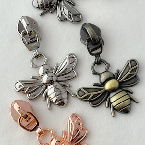 May include: Four zipper pulls with bee charms. The bees are in different finishes: black, silver, gold, and rose gold.
