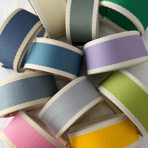 May include: A collection of eleven rolls of grosgrain ribbon in various solid colors including yellow, pink, light blue, dark blue, grey, lavender, lime green, forest green, sage green, and cream, each featuring a contrasting cream-colored border.