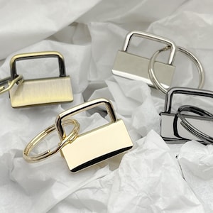 May include: Four key rings with different finishes: gold, silver, black, and brass. Each key ring has a rectangular tag with a smooth surface.