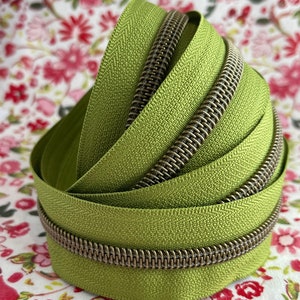 Op de afbeelding: Close-up van een groene ritsband met een messing rits-schuif. De ritsband is gevouwen in een spiraalvorm en ligt op een stoffen achtergrond met bloemen.