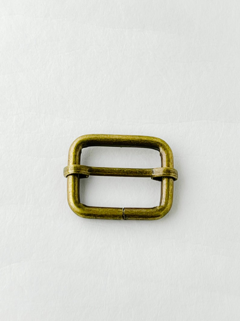 May include: A gold-toned metal buckle with a rectangular shape. The buckle has a smooth, polished finish.