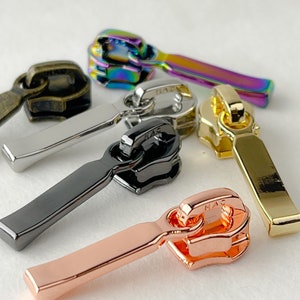 May include: Five zipper pulls in different colors and finishes. The pulls are made of metal and have a simple, rectangular design. The colors are black, silver, gold, rose gold, and iridescent.